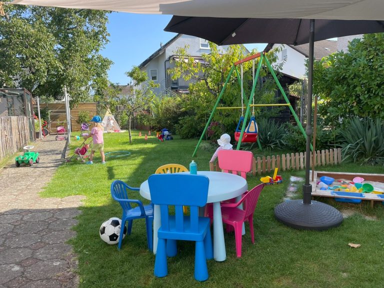 Kindertagespflege Rodenbach - unser Garten Buntes Spielbereich im Garten mit kleinen Möbeln und einem Kind an einem Tisch.