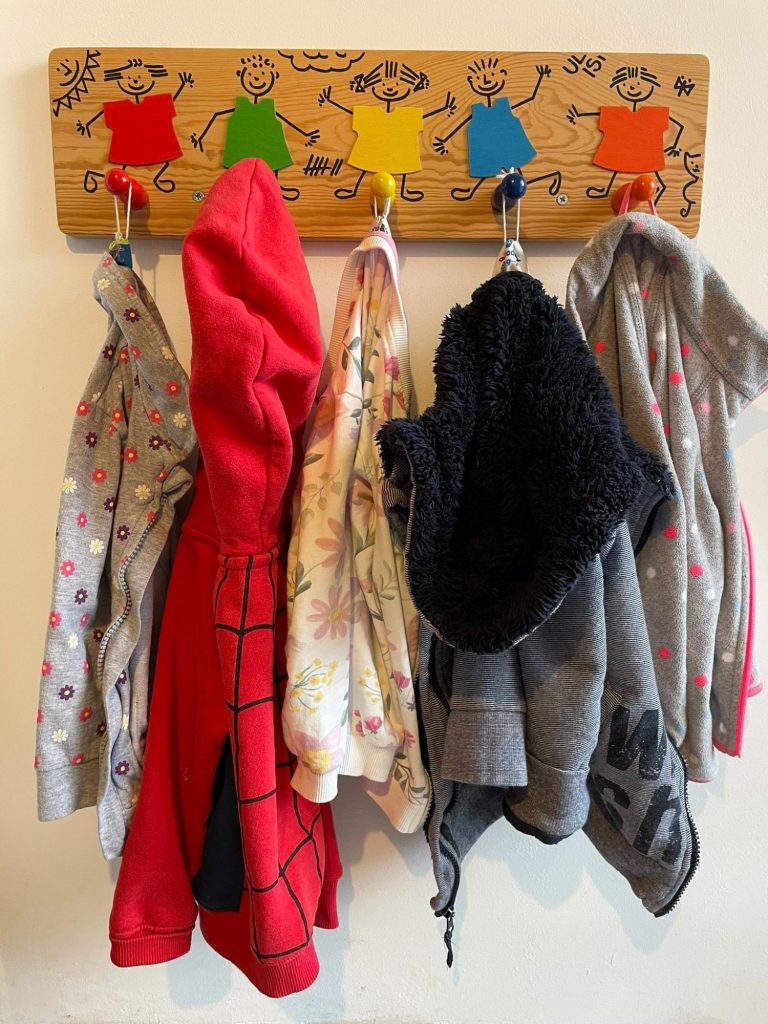 Kindertagespflege Rodenbach - unsere Einrichtung Kleiderhaken mit verschiedenen bunten Kleidungsstücken und einem roten Kapuzenpulli.