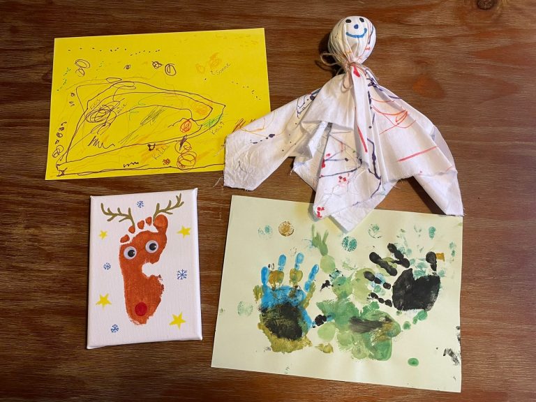 Kindertagespflege Rodenbach - Bastelspaß Bunte Bastelarbeiten: Zeichnungen, Handabdrücke und eine Stoffpuppe auf einem Tisch.